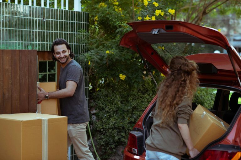 A man and a woman, recently approved for a TN visa, are unloading cardboard boxes from a red car parked near a garden with yellow flowers