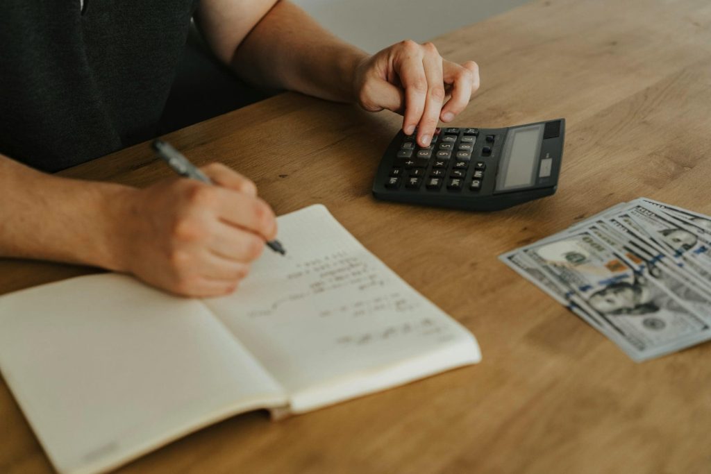 A person uses a calculator and writes in a notebook on a wooden table, with several US hundred-dollar bills placed nearby calculating for estimated taxes.